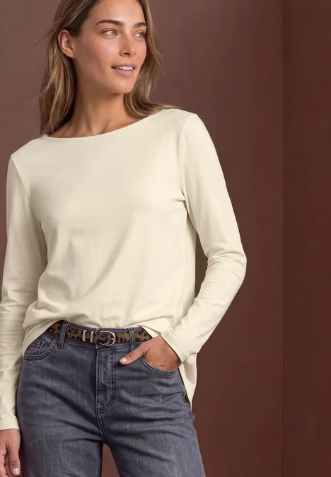 Une femme portant un t-shirt crème à manches longues et un jean, posture décontractée.