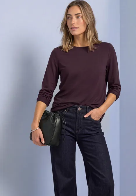 Femme en jean bleu foncé et en haut bordeaux à manches longues, tenant une pochette noire sur fond clair.