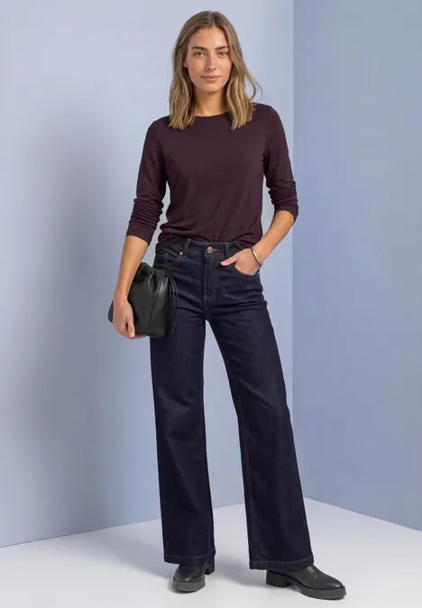 Femme en pull bordeaux, jean large noir et chaussures noires, sac épaule, devant un mur gris-violet.