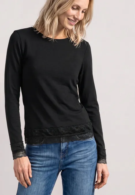 T-shirt à manches longues, col bateau et dentelle Black Frau in schwarzem Langarmshirt mit Spitzenbesatz an Bündchen und Saum, dazu Blue Jeans, lächelnd