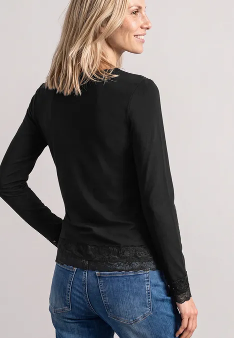 T-shirt à manches longues, col bateau et dentelle Black Frau von hinten in schwarzem Langarmshirt mit Spitzenbund und blauen Jeans, blondes Haar, lächelnd.