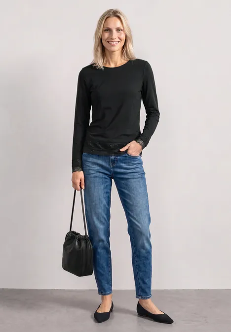 T-shirt à manches longues, col bateau et dentelle Black Frau steht vor hellgrauem Hintergrund, trägt schwarzen Pullover, Jeans, flache Schuhe und hält eine schwarze Tasche.