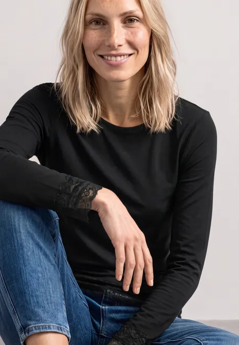 T-shirt à manches longues, col bateau et dentelle Black Eine blonde Frau sitzt lächelnd trägt schwarzen Langarmshirt mit Spitzendetails und blaue Jeans vor schlichten Hintergrund.