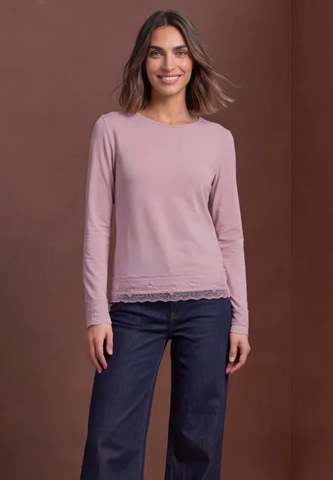 Femme souriante en haut rose à manches longues avec dentelle et jeans bleus devant un fond marron.