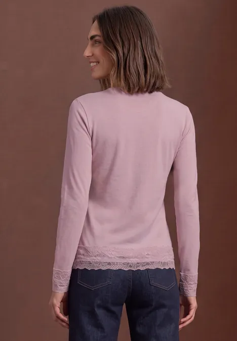 Une femme vue de dos porte un top rose à manches longues avec dentelle et un jean devant un mur brun.