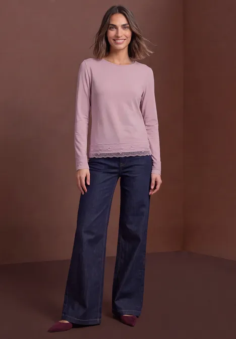 Femme en top rose à manches longues avec dentelle, jean large et chaussures rouges devant un fond marron.