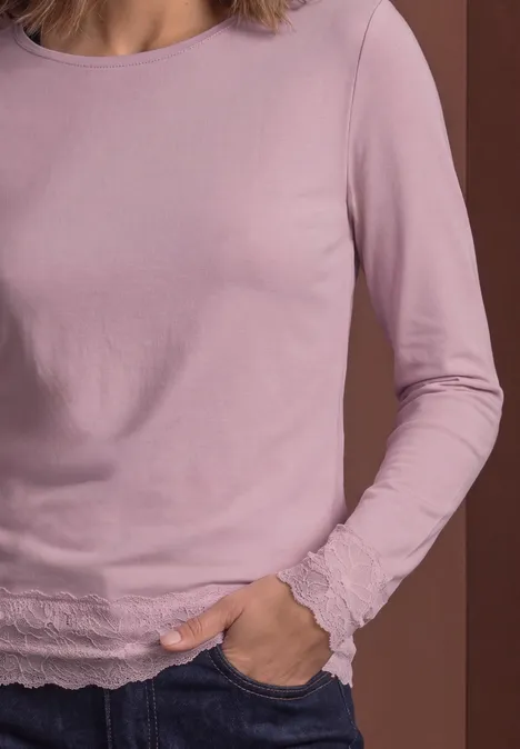 Femme porte un t-shirt à manches longues violet clair avec bordure en dentelle, main dans la poche.