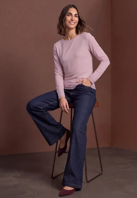 Une femme assise sur un tabouret haut devant un fond brun, en top rose manches longues, jean large et bottines marron.