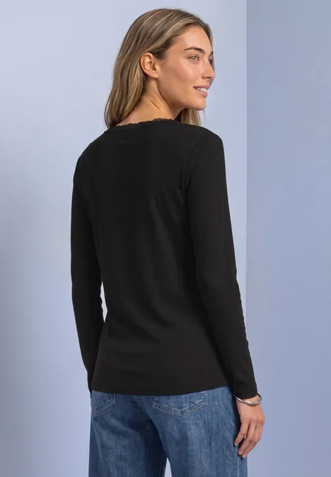 Jasje met fijne ribbel, V-hals en kant Black Profiel van een vrouw in een zwart langarmshirt en blauwe spijkerbroek.