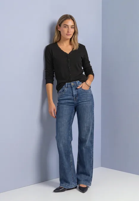Jasje met fijne ribbel, V-hals en kant Black Een vrouw staat nonchalant voor een lichte muur, met een zwarte trui en rechte blauwe jeans.
