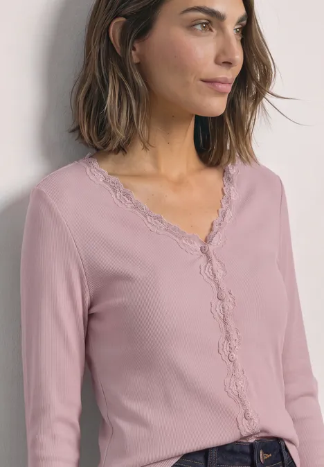 Jasje met fijne ribbel, V-hals en kant quiet rose Frau mit schulterlangem braunem Haar, die ein hellrosa Spitzen-Cardigan mit V-Ausschnitt trägt und seitlich blickt.