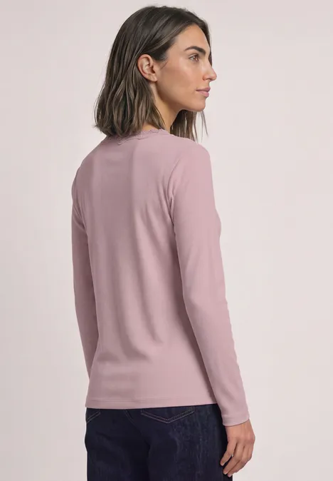 Jasje met fijne ribbel, V-hals en kant quiet rose Eine Frau von hinten in seitlicher Profilansicht trägt ein rosa Langarmshirt und dunkle Jeans vor hellem Hintergrund.