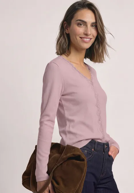 Jasje met fijne ribbel, V-hals en kant quiet rose Frau in rosa Langarmshirt, Jeans und brauner Tasche vor hellem Hintergrund.