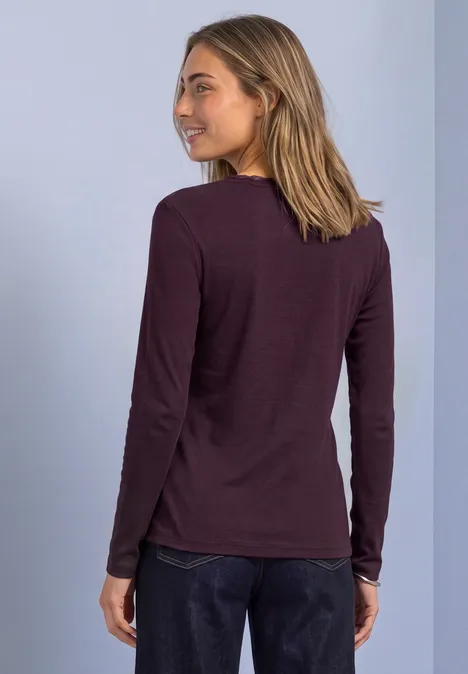 Jasje met fijne ribbel, V-hals en kant berry plum Een vrouw in een donkerpaars shirt met lange mouwen kijkt glimlachend over haar schouder voor een lichtblauwe achtergrond.