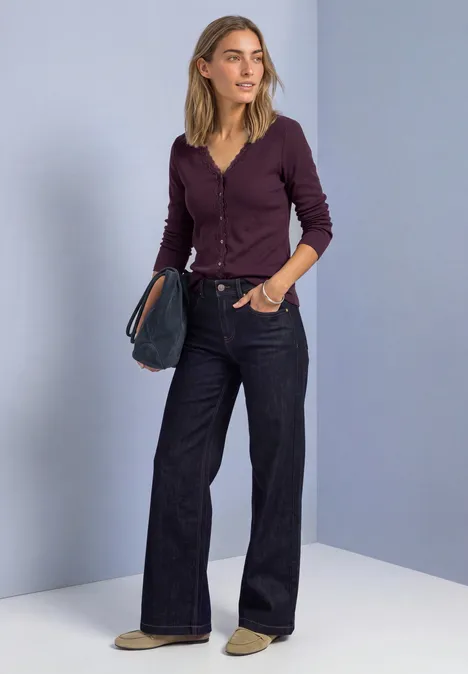 Jasje met fijne ribbel, V-hals en kant berry plum Een vrouw staat voor een lichtpaarse wand, draagt een bordeaux cardigan, wijde jeans en houdt een clutch vast.