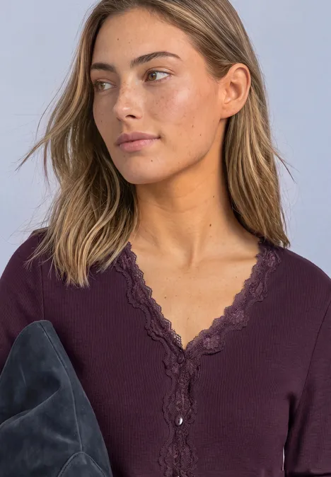 Jasje met fijne ribbel, V-hals en kant berry plum Vrouw met bruin haar in bordeaux top met kant, houdt grijs jasje vast en kijkt opzij.