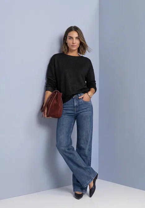 Shirt met gebreide look en vleermuismouwen Black Vrouw voor lichtgrijze muur, zwarte trui, rechte blauwe jeans, zwarte pumps en een bruine handtas.