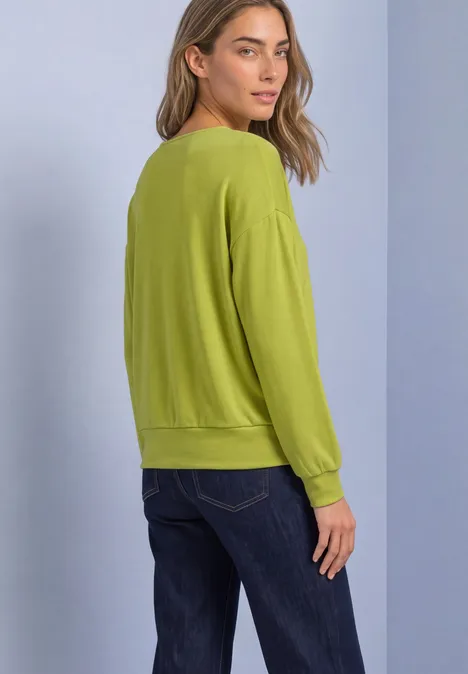 Shirt met gebreide look en vleermuismouwen apple lime Vrouw in profiel met groene trui en blauwe jeans voor een lichte achtergrond.