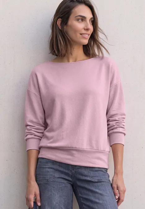 Frau steht lässig vor Wand, trägt rosa Sweatshirt und Jeans, Blick seitlich, entspannte Pose.
