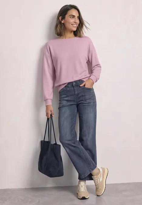 Frau in rosa Pullover, geraden Jeans und Turnschuhen trägt eine blaue Tasche und lächelt.