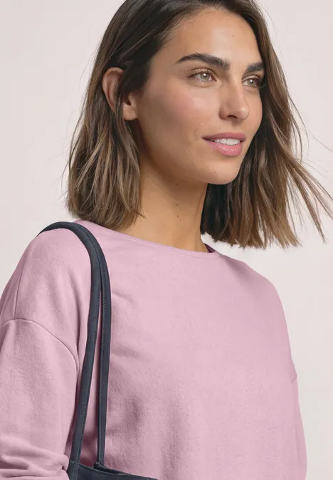 Eine junge Frau mit kurzen braunen Haaren, trägt ein rosa Shirt und eine Schultertasche.