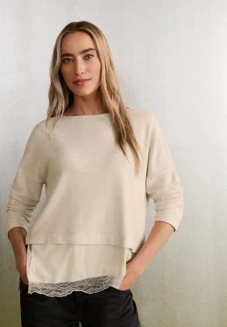 Shirt mit angeschrägter Spitzenkante clay sand mel. Frau mit langen blonden Haaren in beigem Pullover mit Spitzenbesatz, vor neutralem Hintergrund.