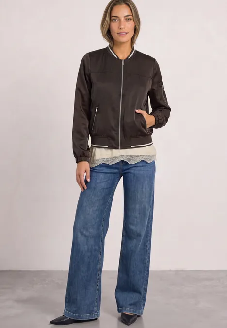 Shirt mit angeschrägter Spitzenkante clay sand mel. Frau trägt schwarze Bomberjacke, weißes Shirt und weite Jeanshose vor neutralem Hintergrund.
