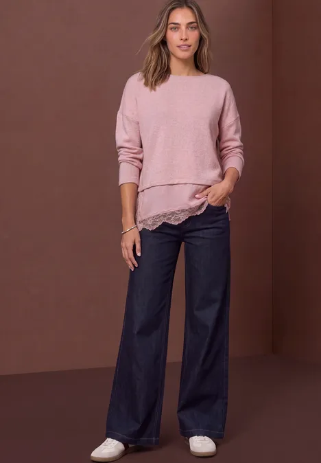 T-shirt avec bordure en dentelle biseautée quiet rose mel. Frau in rosafarbenem Pullover mit Spitzenbesatz, weiten Jeanshosen und weißen Sneakern vor braunem Hintergrund.