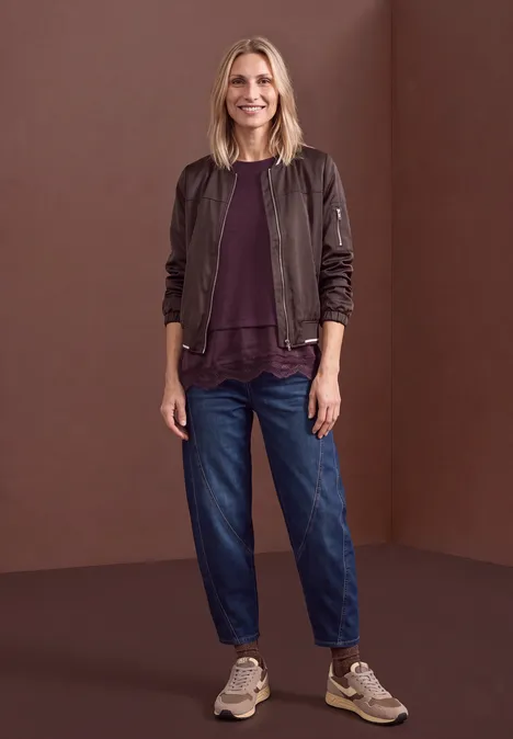 T-shirt avec bordure en dentelle biseautée berry plum mel. Eine lächelnde Frau in Bomberjacke, Jeans und Sneakers vor einer schlichten Wand.