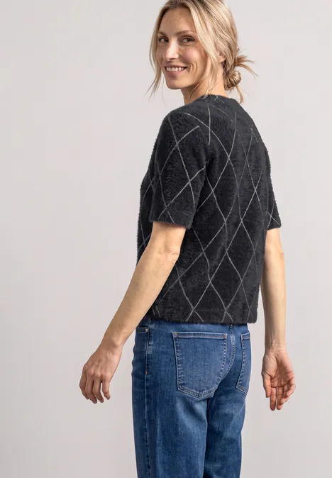 Eine Frau steht seitlich zur Kamera, trägt ein schwarzes, kurzärmeliges Strickoberteil mit Rautenmuster und blaue Jeans.
