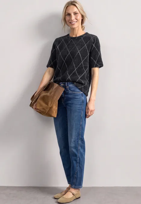 Eine Frau trägt ein schwarzes Shirt mit Karomuster, blaue Jeans, beige Schuhe und eine braune Tasche.