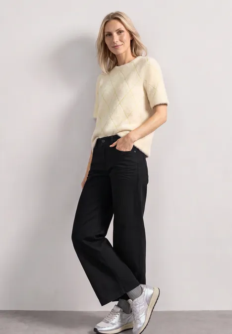 Frau mit kurzem gelbem Pullover, schwarzen weiten Hosen und weißen Sneakers steht vor hellgrauem Hintergrund.