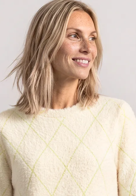 Lächelnde Frau mit schulterlangen blonden Haaren und hellem, kariertem Pullover mit Rautenmuster.