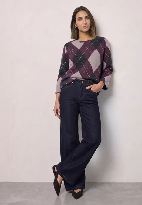Frau in Pullover mit Rautenmuster und weiten Jeans steht vor hellem Hintergrund.
