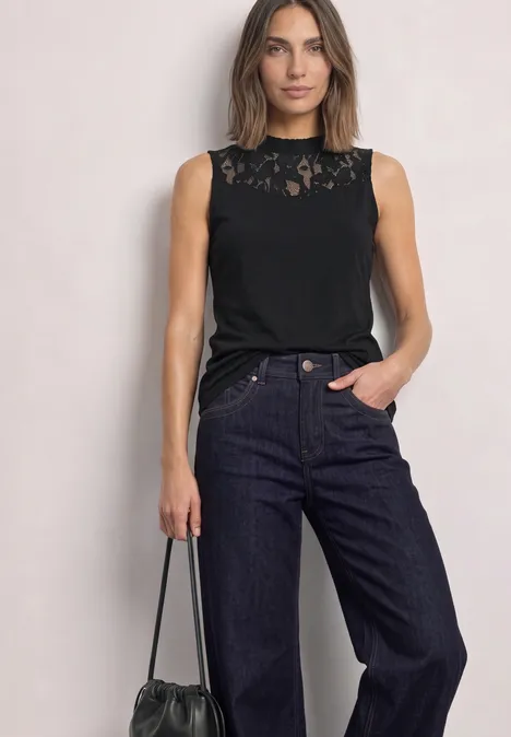Eine Frau trägt ein ärmelloses Spitzen-Top in Schwarz, weite Jeans und hält eine schwarze Tasche.
