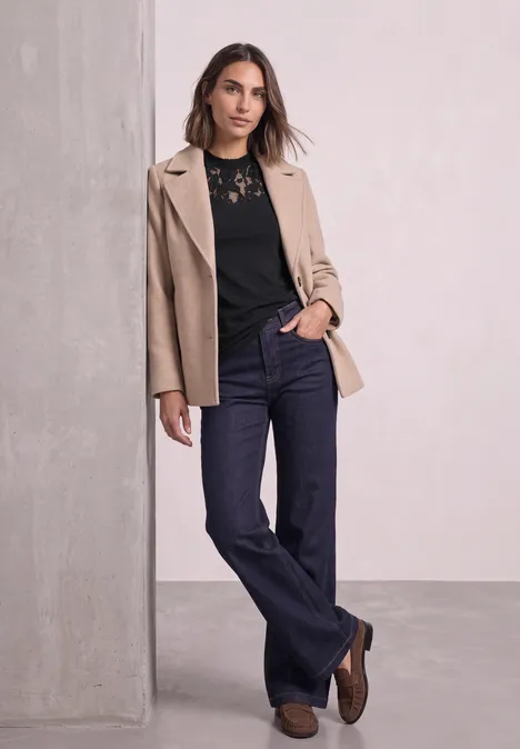 Eine Frau lehnt an einer Säule, trägt beige Blazer, schwarze Spitzenbluse und dunkle Jeans.