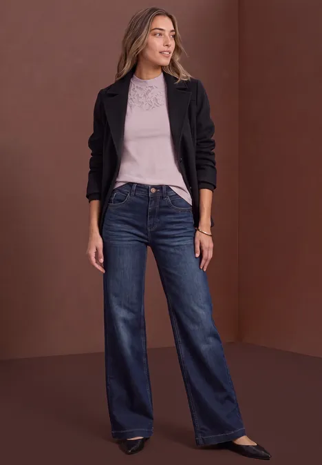 Eine Frau in schwarzer Jacke, grauem Shirt und weiten Jeans posiert vor braunem Hintergrund.