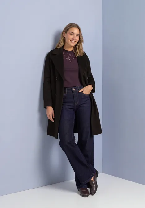 Top mit Rundhals und Spitze berry plum Frau lehnt an pastellblauer Wand, trägt schwarzen Mantel, violettes Oberteil, weite Jeans und braune Schuhe.