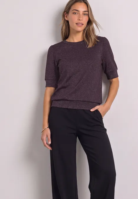 T-Shirt mit Rundhals und Glitzer berry plum Frau trägt violettes Halbarm-Oberteil und schwarze, weite Hose, Hand in Tasche, helle Kulisse.