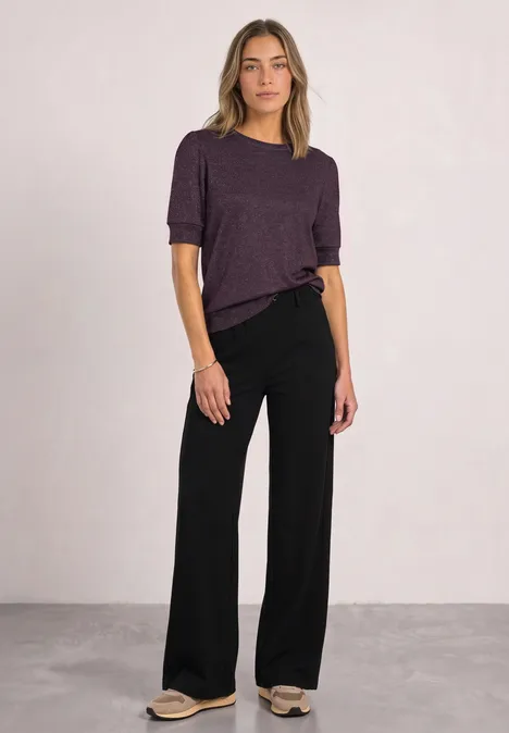 T-Shirt mit Rundhals und Glitzer berry plum Frau steht vor neutralem Hintergrund, trägt lila Oberteil und schwarze, weite Hose.