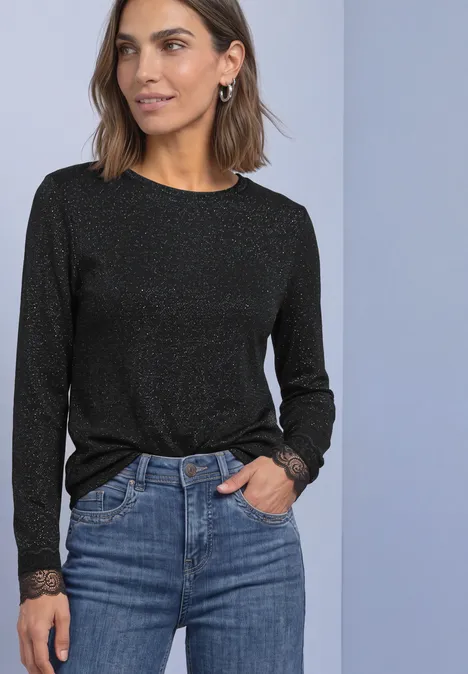 Frau trägt schwarzen, schimmernden Langarmpullover mit Spitzenabschluss an Ärmeln, kombiniert mit Jeans.
