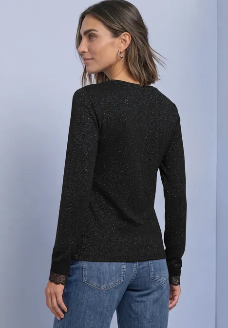Frau von hinten in schwarzem, glitzerndem Langarmshirt und blauer Jeanshose vor einfarbigem Hintergrund.
