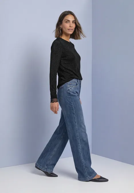 Frau in schwarzem Langarmshirt und weiten blauen Jeans, seitlich stehend auf einfarbigem Hintergrund.