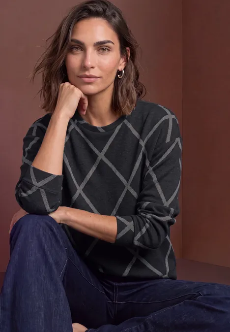 Femme portant un pull sombre à motifs géométriques, assise de manière décontractée devant un fond neutre.