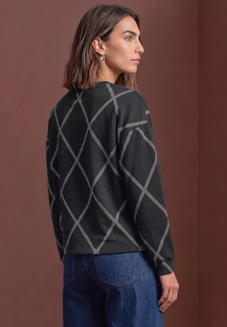 Femme de profil portant un pull noir à losanges gris et un jean bleu.