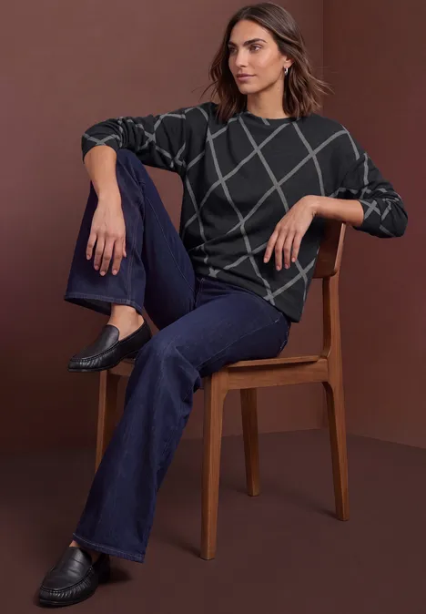 Femme assise sur une chaise, porte un pull gris foncé à motifs quadrillés blancs, jean bleu foncé et mocassins noirs.
