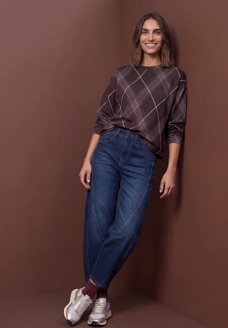 Femme avec un pull à carreaux, jean bleu et baskets blanches devant un fond marron, souriante et décontractée.