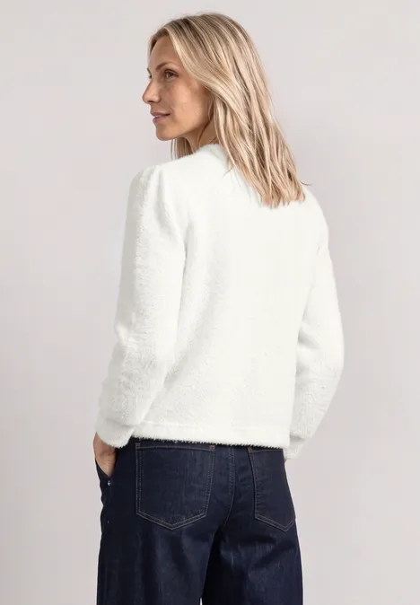 Une femme blonde de dos en veste blanche et jeans bleu sur fond clair.