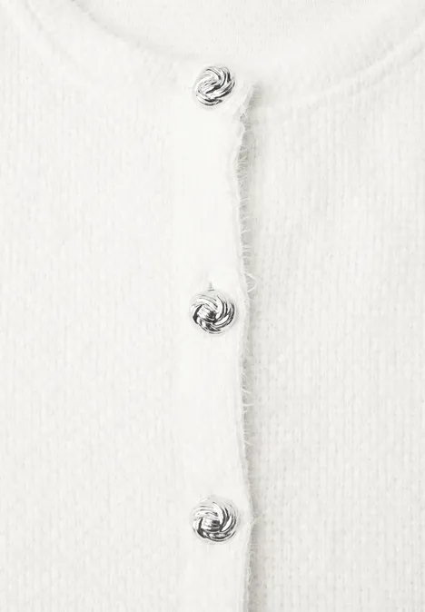 Gros plan d’un cardigan blanc en maille texturée avec des boutons décoratifs argentés alignés sur le devant.