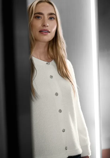 Une femme aux cheveux blonds porte un cardigan blanc en maille et se tient devant un miroir.